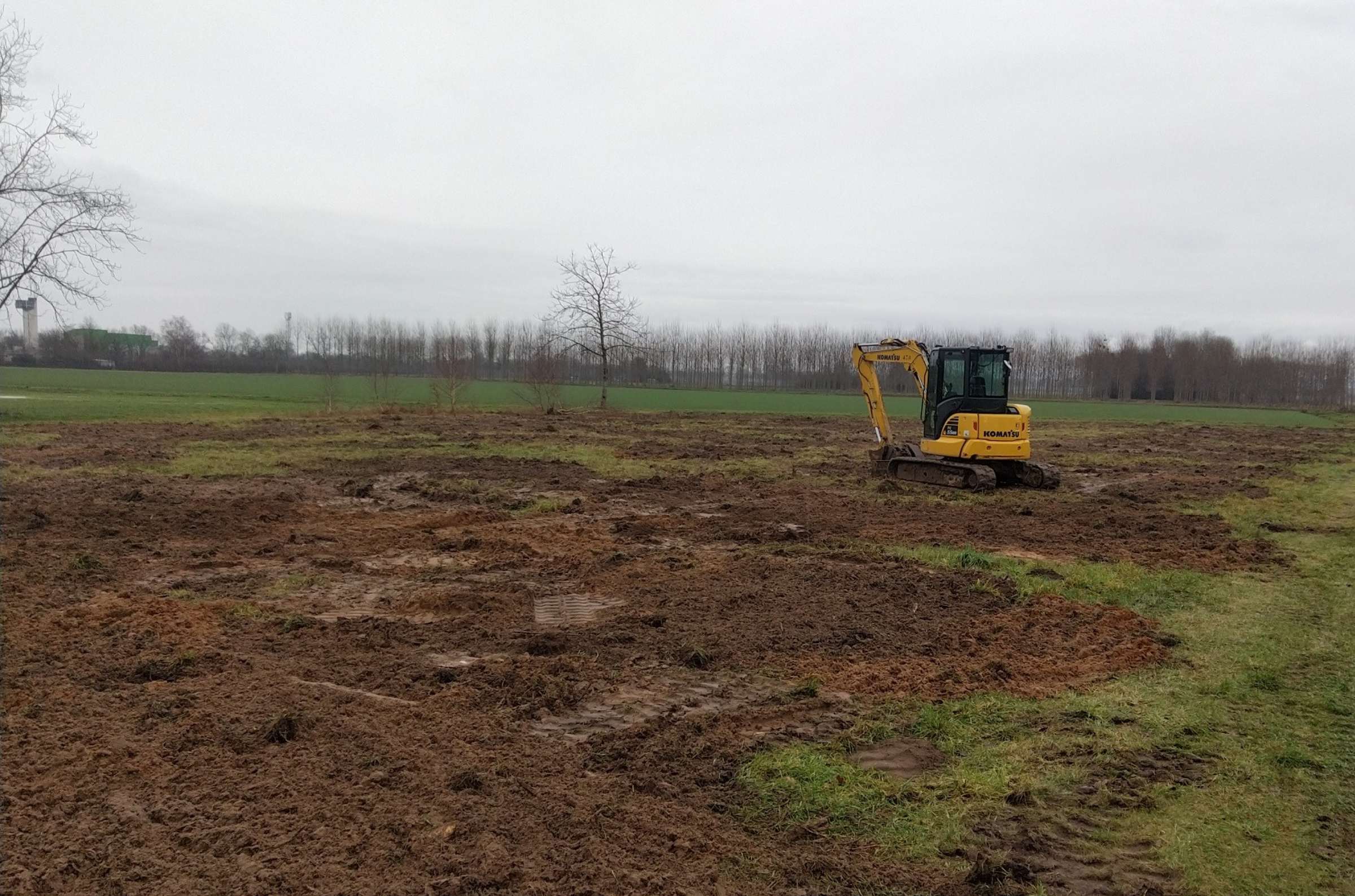 Mini-pelle sur un terrain agricole nu et boueux, prêt pour des travaux de terrassement, sous un ciel couvert.